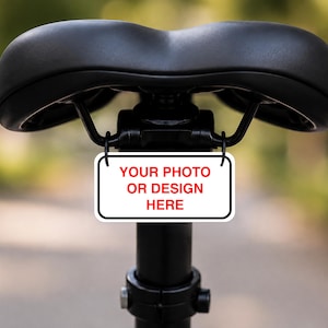 Puede incluir: Un sillín de bicicleta negro montado en una tija con un pequeño letrero rectangular blanco colgado de los raíles. El letrero tiene un borde negro y el texto rojo "YOUR PHOTO OR DESIGN HERE" impreso en el centro de la superficie blanca.