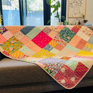 May include: A patchwork quilt featuring a mix of floral and geometric patterns in pink, yellow, green, blue, and white. The quilt is folded and draped over a grey sofa.