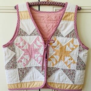 May include: A patchwork quilted vest with star patterns in pink and yellow. The vest has a white background with floral and geometric accents, and is trimmed with a red and white gingham pattern. It is hanging on a vintage hanger.