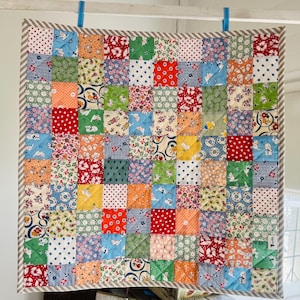 May include: A patchwork quilt featuring a vibrant mix of patterned squares in shades of red, blue, green, orange, and yellow. The quilt is edged with a striped fabric and is suspended by blue clips. The quilt is square in shape.