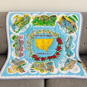 May include: A light blue souvenir tea towel featuring a map of New South Wales and Canberra, Australia. The towel has a white border with a purple and white gingham trim. The map is surrounded by illustrations of landmarks and the text "NEW SOUTH WALES" and "CANBERRA A.C.T."