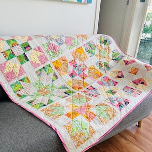 May include: A colourful patchwork quilt with floral and geometric patterns in pink, green, yellow, and orange. The quilt has a white background and a pink and white striped border. It is draped over a grey sofa. The quilt's dimensions are not visible.
