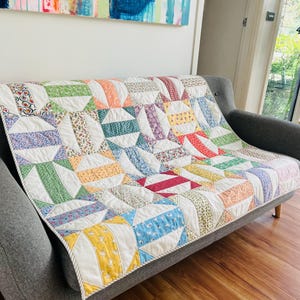 May include: A patchwork quilt in a variety of colours, draped over a grey sofa. The quilt has a repeating pattern of curved shapes and stripes in shades of white, green, yellow, blue, and red. A decorative home textile.