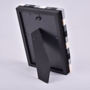 May include: Black picture frame with a white and black checkered pattern on the border. The frame has a stand on the back.