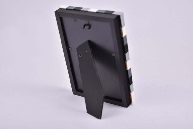 May include: Black picture frame with a white and black checkered pattern on the border. The frame has a stand on the back.