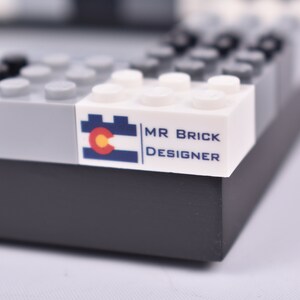 May include: A white LEGO brick with a blue and red Colorado flag and the text "MR BRICK DESIGNER" printed on it.