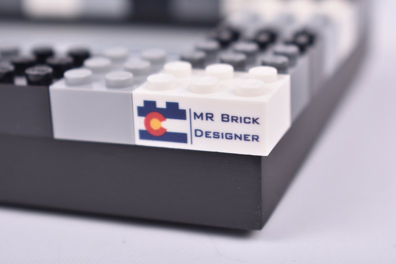 May include: A white LEGO brick with a blue and red Colorado flag and the text "MR BRICK DESIGNER" printed on it.