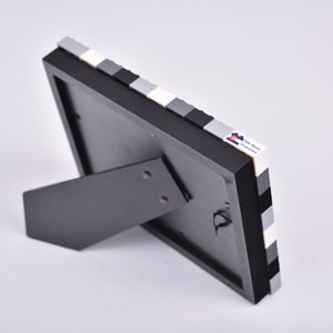 May include: Black picture frame with a white and gray Lego brick border. The frame has a kickstand on the back.