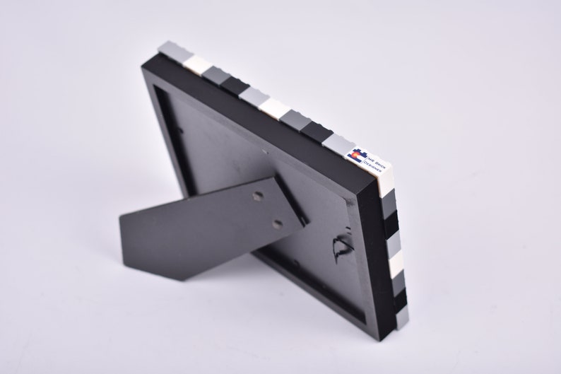 May include: Black picture frame with a white and gray Lego brick border. The frame has a kickstand on the back.