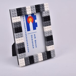 May include: A black, white, and gray Lego picture frame with a business card inside. The business card has the text "MR BRICK DESIGNER" and "www.mrbrickdesigner.com".