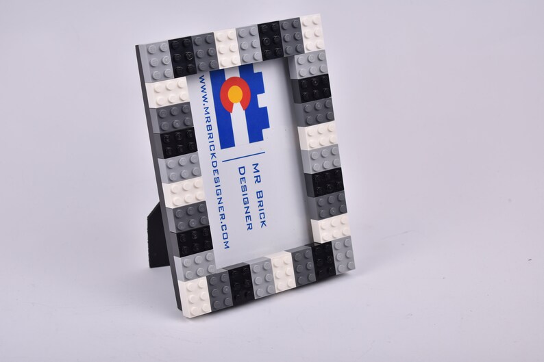 May include: A black, white, and gray Lego picture frame with a business card inside. The business card has the text "MR BRICK DESIGNER" and "www.mrbrickdesigner.com".