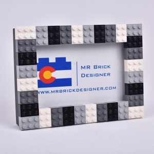 May include: A black, white, and gray Lego picture frame with a business card inside. The business card says "Mr. Brick Designer" and "www.mrbrickdesigner.com".