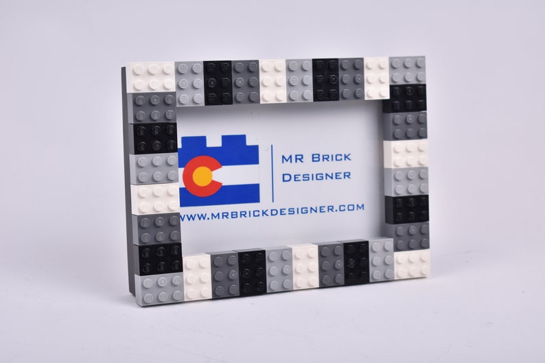 May include: A black, white, and gray Lego picture frame with a business card inside. The business card says "Mr. Brick Designer" and "www.mrbrickdesigner.com".