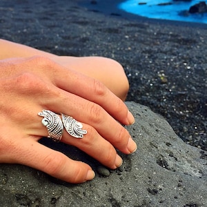 May include: A silver ring with a detailed design resembling wings. The ring is worn on a person's finger, resting on a dark gray rock.