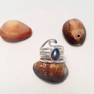 May include: A silver ring with a wave design and a dark gray pearl. The ring is sitting on a seashell.