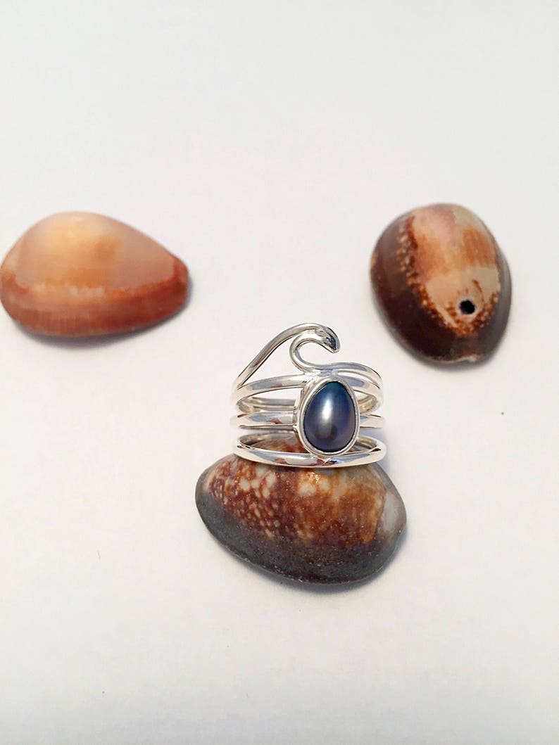 May include: A silver ring with a wave design and a dark gray pearl. The ring is sitting on a seashell.