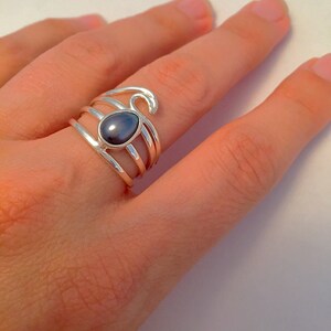 May include: A silver ring with a dark gray oval gemstone set in the center. The ring has a wave design on the band.