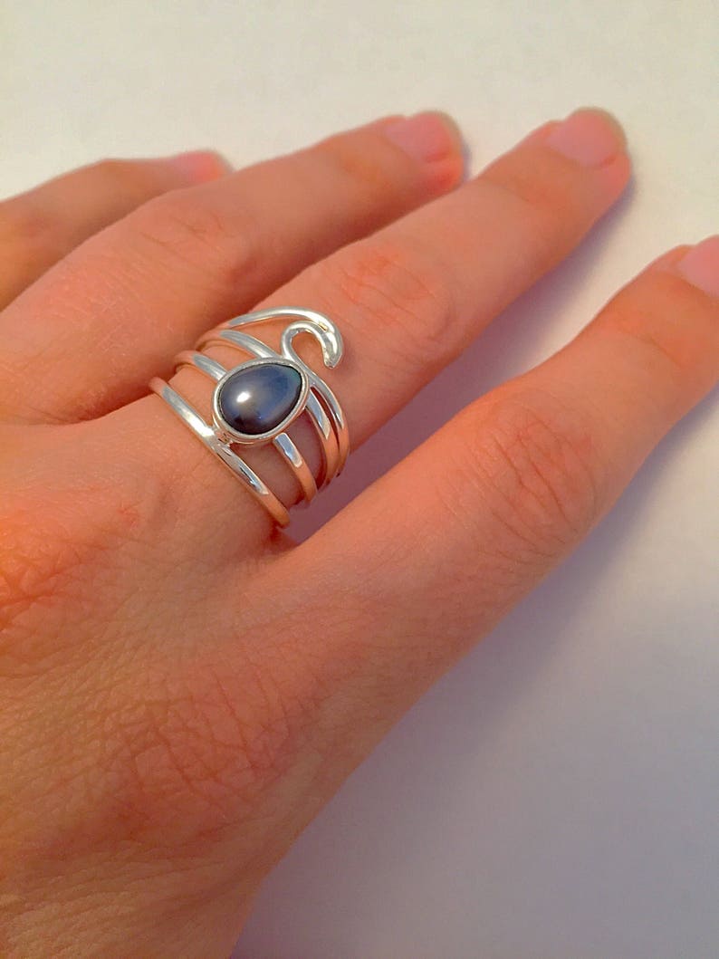 May include: A silver ring with a dark gray oval gemstone set in the center. The ring has a wave design on the band.