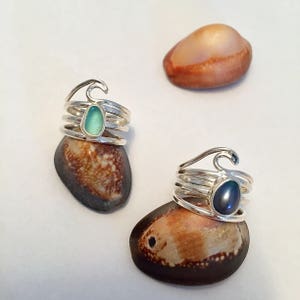 May include: Two silver rings with a wave design. One ring has a light blue stone and the other has a dark blue stone.