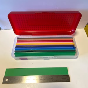 May include: A red plastic case with an open lid, revealing a clear container filled with colorful long strips. A green strip and a metal ruler are below the case. The ruler is marked with inches.