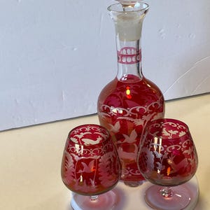 May include: A cranberry red glass decanter with a matching stopper and two matching snifter glasses. Each piece features intricate white etched designs of birds, deer, and floral patterns. The glass has a deep red hue with clear glass bases.
