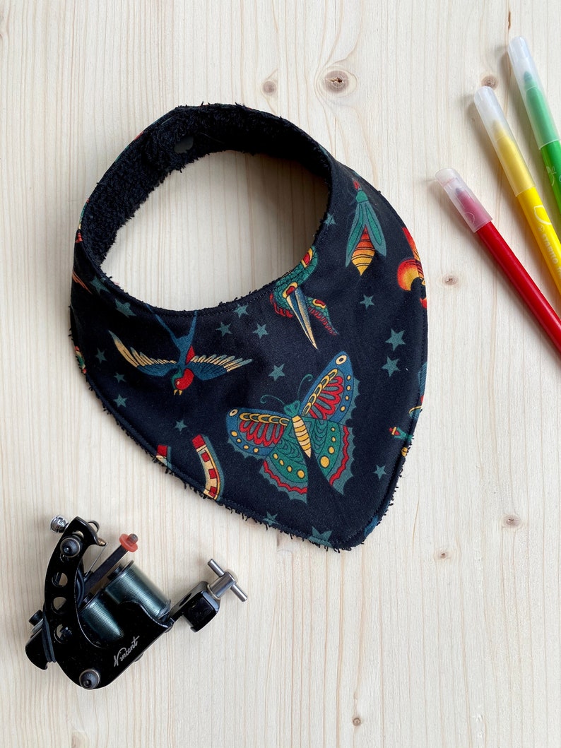K&ouml;nnte beinhalten: Ein schwarzes Bandana mit einem wei&szlig;en Stern und bunten Tattoo-Designs von einem Schmetterling, einem Vogel und einem Insekt. Das Bandana hat einen schwarzen Frottee-Rand.