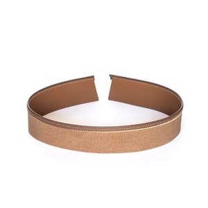May include: A brown headband with a textured, glittery surface. The headband has a stitched edge and a gap in the center. The headband is made of a flexible material.