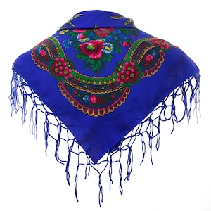 Può includere: Un foulard quadrato blu con un motivo floreale e frange. Il foulard presenta un design floreale colorato con fiori rossi, gialli e verdi.
