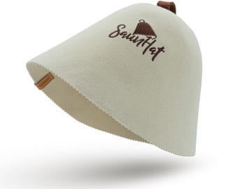 Puede incluir: Un gorro de sauna de fieltro blanco con una correa de cuero marrón y el texto "Saunhat" bordado en la parte delantera.