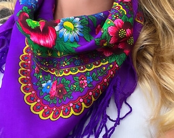 May include: A purple paisley patterned scarf with a fringe border. The scarf is tied around the neck of a person with blonde hair.