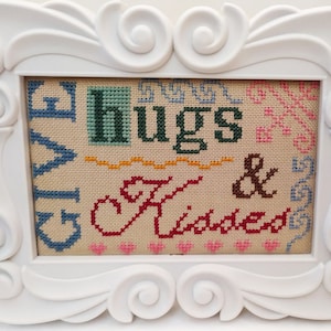 Pode incluir: Uma moldura branca com um design de ponto cruz colorido que diz "Give Hugs & Kisses".
