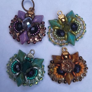 Może przedstawiać: Cztery koraliki w kształcie sowy w różnych schematach kolorystycznych. Koraliki są wykonane z koralików nasiennych, koralików szklanych i metalowych okuć. Koraliki są w odcieniach fioletu, turkusu, zieleni i brązu.