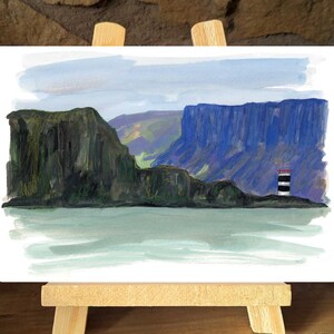 Può includere: Dipinto ad acquerello di un paesaggio costiero con un faro. L'opera raffigura un'isola rocciosa con un faro bianco e nero, sullo sfondo di scogliere blu e un cielo azzurro. Il dipinto è esposto su un piccolo cavalletto di legno.