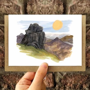 Op de afbeelding: Een aquarel landschaps wenskaart met een berglandschap met een grote, donkere rotsformatie op de voorgrond. De zon is een heldergele cirkel. De kaart wordt tegen een bakstenen achtergrond gehouden.