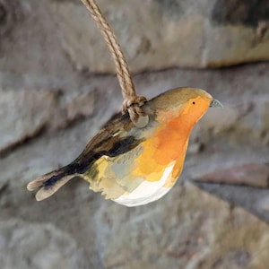 Puede incluir: Una pintura de acuarela de un petirrojo con un esquema de color marrón, naranja y blanco. El pájaro está colgado de una cuerda y mira hacia la derecha. El fondo es una imagen borrosa de una pared de piedra.