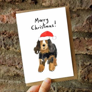 Puede incluir: Una tarjeta de Navidad con una ilustración en acuarela de un perro con un gorro de Papá Noel. La tarjeta tiene el texto "Merry Christmas!" escrito en escritura negra. La tarjeta es blanca con un borde marrón.