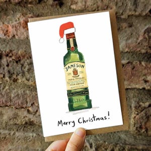 Può includere: Un biglietto di Natale ad acquerello con una bottiglia verde di Jameson Irish Whiskey con un cappello da Babbo Natale. Il biglietto dice "Merry Christmas!" in un carattere scritto a mano. Il biglietto è su uno sfondo marrone.