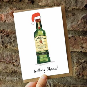 Può includere: Biglietto d'auguri natalizio con un'illustrazione ad acquerello di una bottiglia di whisky irlandese Jameson con un cappello da Babbo Natale. Il biglietto include il testo "Nollaig Shona!", un saluto festoso irlandese.
