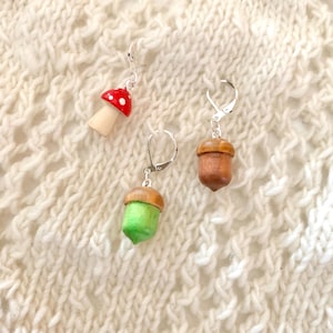 May include: Three pairs of earrings with nature-inspired designs. One pair features a red mushroom with white spots, and the other two pairs are acorns, one green and one brown. All earrings have silver-coloured leverback closures.