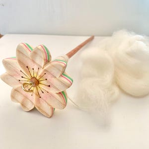 以下が含まれることがあります： ピンク、緑、ベージュのストライプが入った、花型の輪が付いた木製のドロップスピンドル。 スピンドルは長い茶色のシャフトと金色のフックが付いています。 その隣には、ふわふわの白いウールロービングが2つあります。