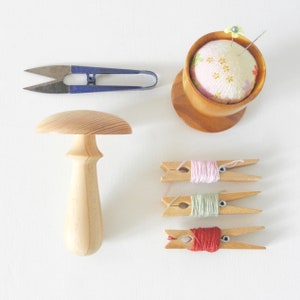 May include: A selection of sewing items, including blue-handled thread snips, a wooden mushroom-shaped thread holder, a pincushion, and three clothespins with coloured thread spools. The items are arranged on a white surface.