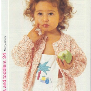 May include: A young child wearing a pink knitted cardigan with a white button-up shirt and a white onesie with a colourful embroidered design. The child is holding a green apple.