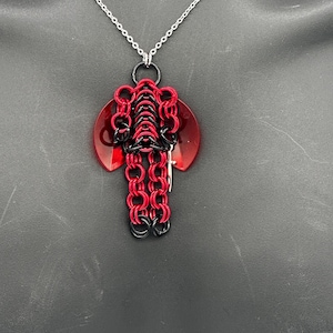 May include: A silver chain necklace with a pendant featuring a red and black chainmail design. The pendant has a red, translucent, shield-shaped backing. The chainmail is red and black, with a small silver accent.