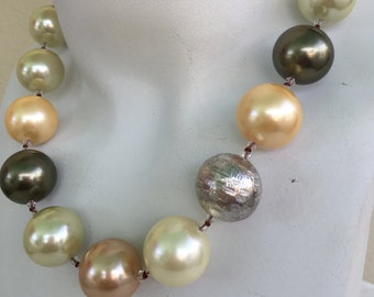 May include: A necklace made of large, round, cream, gold, and green beads. The beads are strung on a thin, brown cord. The necklace is displayed on a white mannequin.