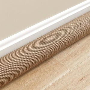 May include: A cylindrical draft stopper featuring a tight, woven fabric pattern in shades of tan and dark brown. The long, tubular object is positioned horizontally along the base of a wall, resting against the white trim and light wood flooring.