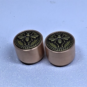 May include: Two gold-coloured metal knobs with a bee design on the top. The bee is black and gold and is surrounded by a gold chain design.