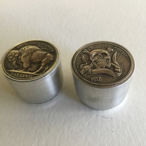 Puede incluir: Dos tapones de botella de forma de moneda de plata. El tapón de la izquierda presenta un búfalo de color dorado con el texto "UNITED STATES OF AMERICA" y "FIVE CENTS". El tapón de la derecha presenta una calavera y huesos cruzados dorados con el texto "LIBERTY" y "1937".
