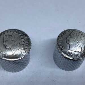 May include: Two silver-colored 1904 Indian Head pennies are mounted on a silver-colored base. The pennies are facing each other.