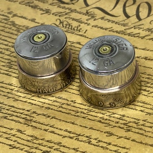 Peut inclure: Deux bagues en argent en forme de douilles de fusil de chasse avec une pièce de monnaie d'un demi-dollar et une pièce de monnaie de 25 cents de 1995. Les bagues sont sur un document jauni avec du texte.