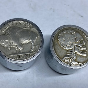 Puede incluir: Dos objetos metálicos de forma redonda de color plateado con un borde elevado. El objeto de la izquierda presenta un búfalo en la parte delantera y el texto "UNITED STATES OF AMERICA" y "FIVE CENTS". El objeto de la derecha presenta una calavera en la parte delantera y el texto "LIBERTY" y "1937".